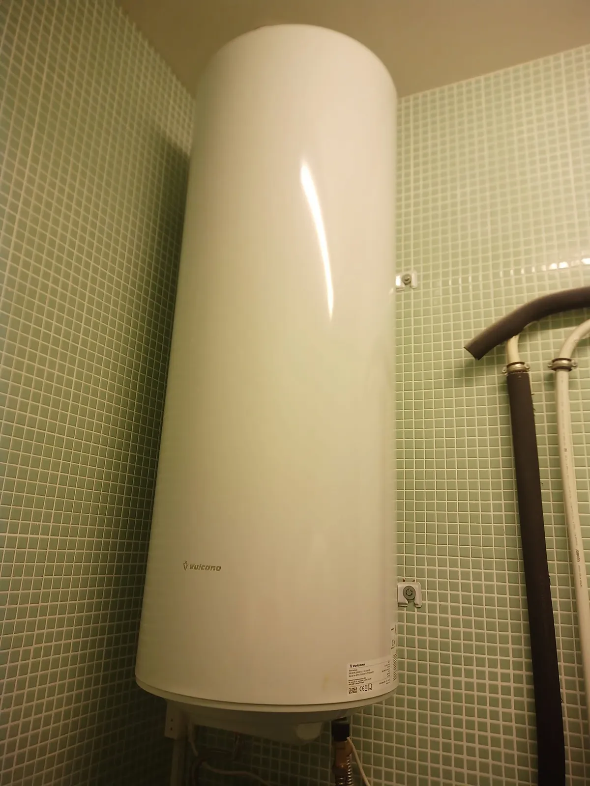 Termoacumulador branco vertical numa parede de pastilha verde, com os tubos ligados