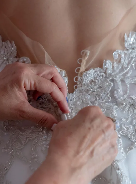 Mãos a abotoar as costas de um vestido de noiva com renda bordada.