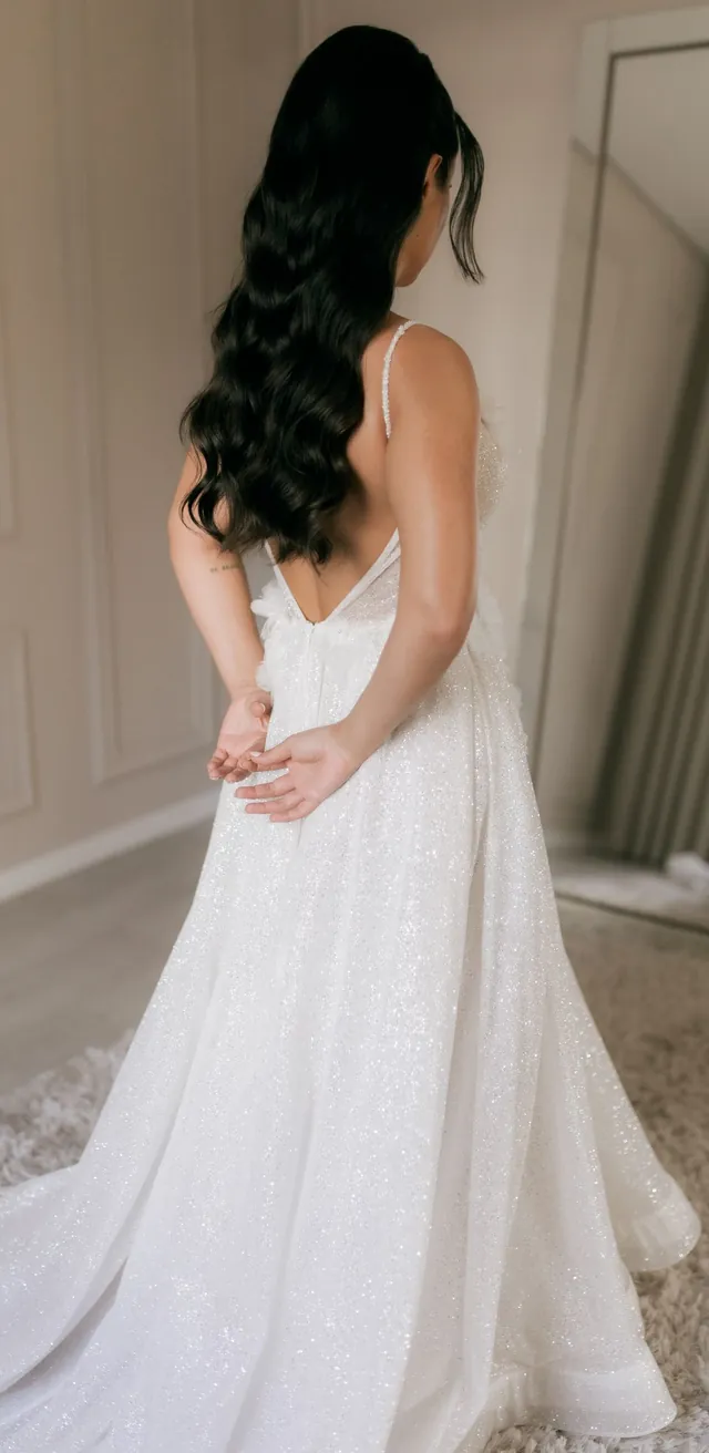 Noiva de costas no atelier, com um vestido branco de alças e cauda comprida.