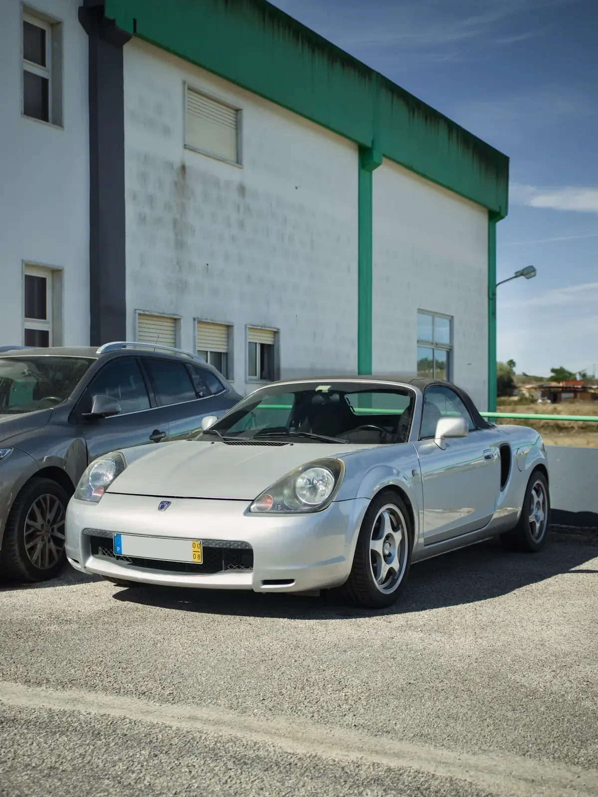 Toyota MR2 descapotável prateado estacionado junto a um pavilhão